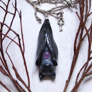 May include: A silver necklace with a branch design and a hanging bat charm. The bat is black with a purple gemstone in its chest. The necklace also has a small silver key charm.