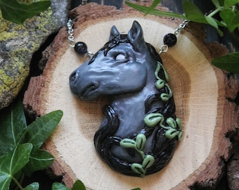 May include: A handcrafted necklace featuring a gray horse head pendant with black mane and green ivy accents. The pendant is on a silver chain with black bead accents, set against a wooden slice and surrounded by green leaves.