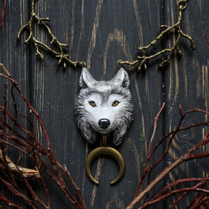 Puede incluir: Un collar con un colgante de cabeza de lobo plateado y un dije de media luna. El collar está hecho de metal y tiene un diseño de rama. El colgante está colgando sobre un fondo de madera oscura.