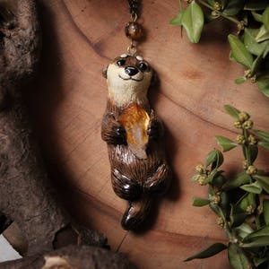May include: A brown and white otter charm necklace with a golden crystal. The otter is holding the crystal in its paws. The necklace is on a wooden surface with green leaves.