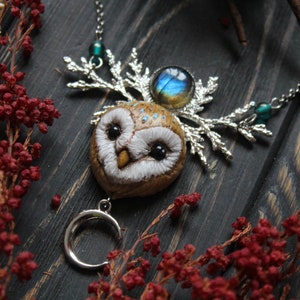 May include: A silver necklace with a brown owl pendant, a silver crescent moon charm, and a blue iridescent gemstone. The owl has white feathers and black eyes. The necklace is on a dark wooden background with red berries.