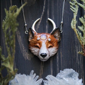 May include: A silver chain necklace with a fox head pendant. The fox is made of brown and white clay and has a crescent moon above its head. The fox has three white stones on its forehead.
