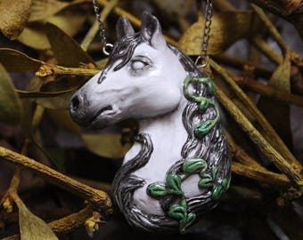 May include: A white ceramic horse head pendant necklace with green leaves and vines. The necklace is hanging from a silver chain against a background of dried leaves.