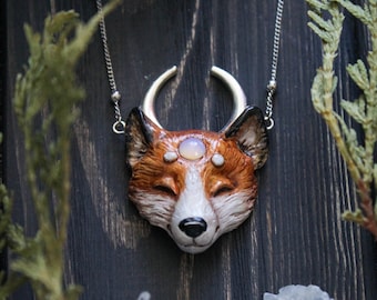 May include: A silver chain necklace with a fox head pendant. The fox is made of brown and white clay and has a crescent moon above its head. The fox has three white stones on its forehead.