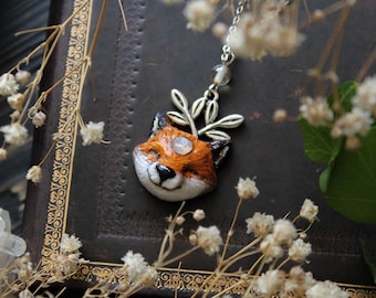 May include: A handcrafted fox pendant necklace. The pendant features a smiling fox head with orange, white, and black details, a gemstone eye, and a silver-colored leaf accent. The necklace is displayed on a dark brown leather-bound book, surrounded by dried flowers.