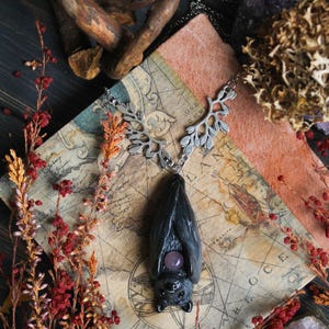 May include: A dark gray bat pendant with a silver-colored leaf design chain and a purple gemstone. The pendant is displayed on a vintage map, surrounded by dried flowers, crystals, and a piece of wood.
