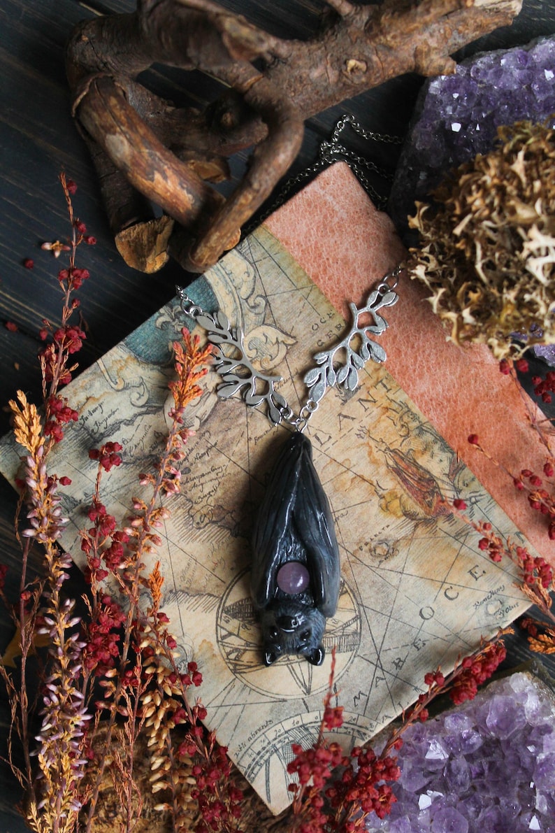 May include: A dark gray bat pendant with a silver-colored leaf design chain and a purple gemstone. The pendant is displayed on a vintage map, surrounded by dried flowers, crystals, and a piece of wood.