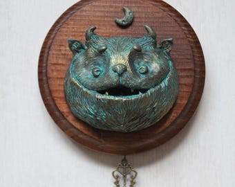 May include: A whimsical wall hanging featuring a smiling creature with horns, a crescent moon, and a decorative key. The creature is teal and gold, mounted on a round wooden plaque. The key hangs below the plaque.