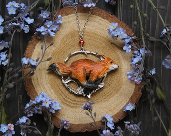 May include: A silver necklace with a fox pendant. The fox is running and is made of painted polymer clay. The necklace is on a wooden surface with blue flowers.