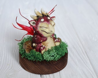 May include: A small, red and white dragon figurine with closed eyes, sitting on a bed of green moss and surrounded by small, clear crystals. The dragon has red wings and is perched on a wooden base.