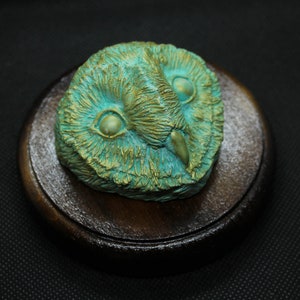 May include: A detailed, teal and gold owl sculpture, featuring intricate feather details and large, expressive eyes. The owl head is mounted on a round, dark wooden base, creating a striking contrast. The sculpture is a decorative item.