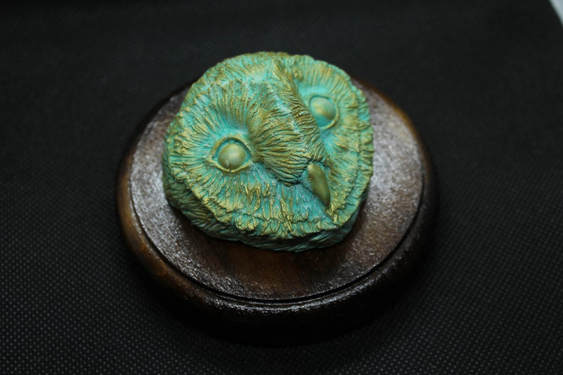 May include: A detailed, teal and gold owl sculpture, featuring intricate feather details and large, expressive eyes. The owl head is mounted on a round, dark wooden base, creating a striking contrast. The sculpture is a decorative item.