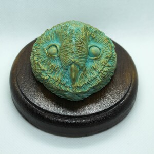 May include: A detailed sculpture of an owl's face, featuring a teal and gold color scheme. The owl is mounted on a round, dark brown wooden base. The sculpture has a textured appearance, with visible feather details.
