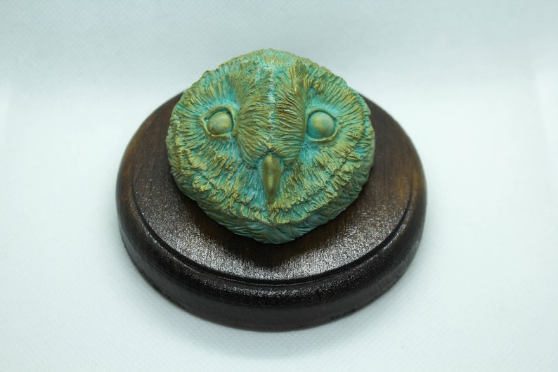 May include: A detailed sculpture of an owl's face, featuring a teal and gold color scheme. The owl is mounted on a round, dark brown wooden base. The sculpture has a textured appearance, with visible feather details.