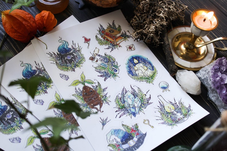May include: A sheet of stickers featuring whimsical illustrations of a cauldron, a potion bottle, a crystal, a plant, a candle, a hat, and a key. The stickers are surrounded by a floral design with lavender flowers and ferns.