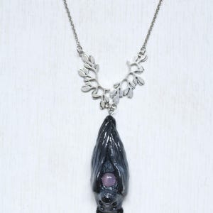 May include: A silver-toned necklace featuring a bat pendant. The bat is dark gray with a purple gemstone belly. The necklace has a decorative leaf-shaped upper chain and a delicate chain.