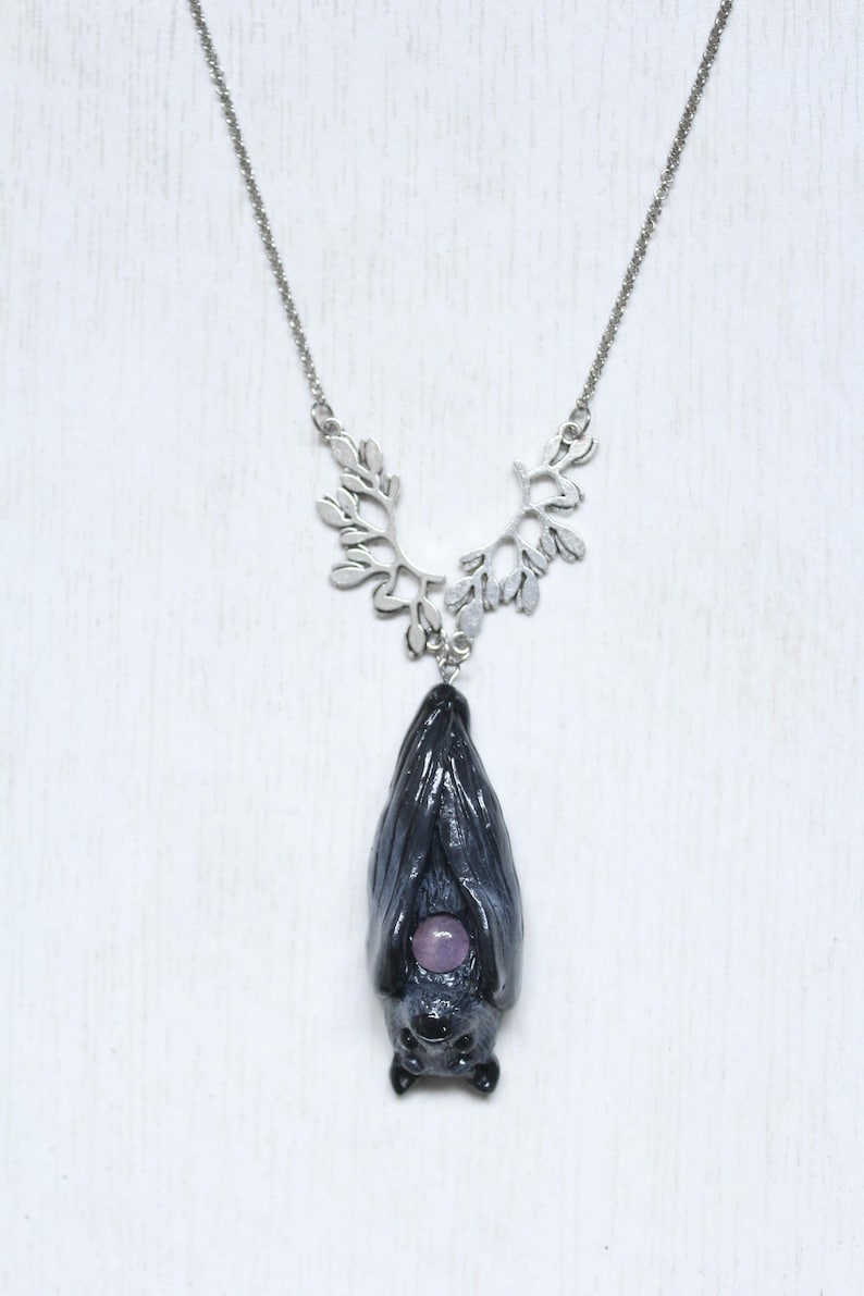 May include: A silver-toned necklace featuring a bat pendant. The bat is dark gray with a purple gemstone belly. The necklace has a decorative leaf-shaped upper chain and a delicate chain.