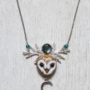 May include: A silver chain necklace with a brown and white owl pendant. The owl has a blue and green gemstone on its head and a silver crescent moon charm hanging below. The necklace is on a white wooden background.