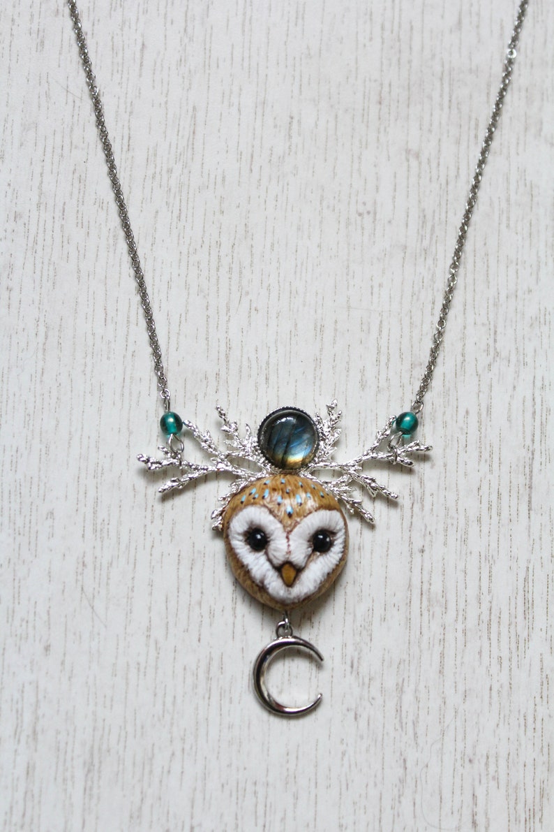 May include: A silver chain necklace with a brown and white owl pendant. The owl has a blue and green gemstone on its head and a silver crescent moon charm hanging below. The necklace is on a white wooden background.