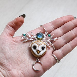 May include: A silver necklace with a felt owl pendant, a labradorite gemstone, and a crescent moon charm. The owl has green eyes and a white face. The necklace is on a hand.