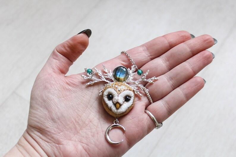 May include: A silver necklace with a felt owl pendant, a labradorite gemstone, and a crescent moon charm. The owl has green eyes and a white face. The necklace is on a hand.