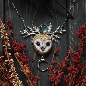 May include: A silver necklace with a white owl pendant, a crescent moon charm, and a labradorite gemstone. The owl has brown and white feathers and black eyes. The necklace is on a dark wooden background with red and white flowers.
