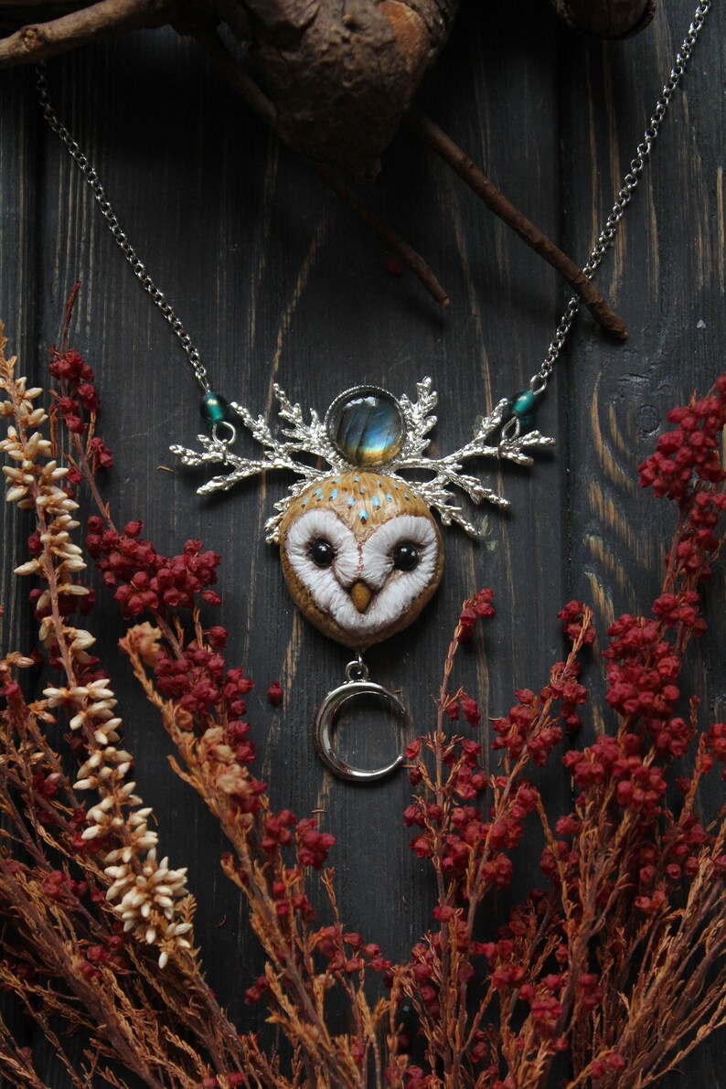 May include: A silver necklace with a white owl pendant, a crescent moon charm, and a labradorite gemstone. The owl has brown and white feathers and black eyes. The necklace is on a dark wooden background with red and white flowers.