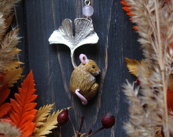 May include: A handcrafted pendant featuring a detailed mouse figurine with a pink tail, suspended from a silver ginkgo leaf charm. The pendant is accented with a small, round, pink gemstone. The background includes dried plants and autumn leaves.