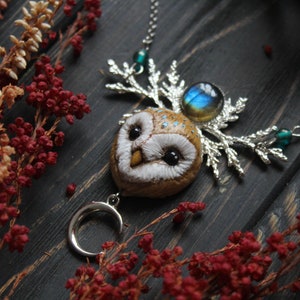May include: A silver necklace with a brown owl pendant, a silver crescent moon charm, and a blue gemstone. The owl has white feathers and black eyes. The necklace is on a dark wooden surface with red and white flowers.