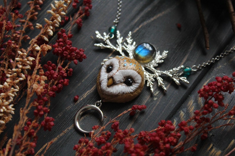 May include: A silver necklace with a brown owl pendant, a silver crescent moon charm, and a blue gemstone. The owl has white feathers and black eyes. The necklace is on a dark wooden surface with red and white flowers.