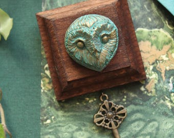 May include: A wooden box with a teal owl face sculpture. A bronze key and a keyhole are also present. The owl sculpture has detailed feather textures. The background is a mix of green and teal.