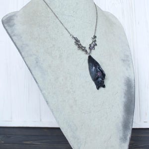 May include: A silver-toned necklace with a bat pendant. The bat pendant is dark gray with a hint of purple and is suspended from a delicate chain. The necklace is displayed on a gray bust.