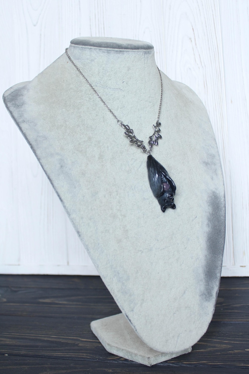 May include: A silver-toned necklace with a bat pendant. The bat pendant is dark gray with a hint of purple and is suspended from a delicate chain. The necklace is displayed on a gray bust.