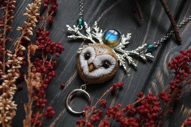 May include: A silver necklace with a brown owl pendant, a silver crescent moon charm, and a blue iridescent gemstone. The owl has white feathers and black eyes. The necklace is on a dark wooden background with red berries.