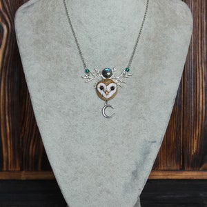 May include: A silver chain necklace with a felt owl pendant. The owl has a brown and white pattern and a green gemstone on its head. The pendant is attached to a silver branch with a crescent moon charm.