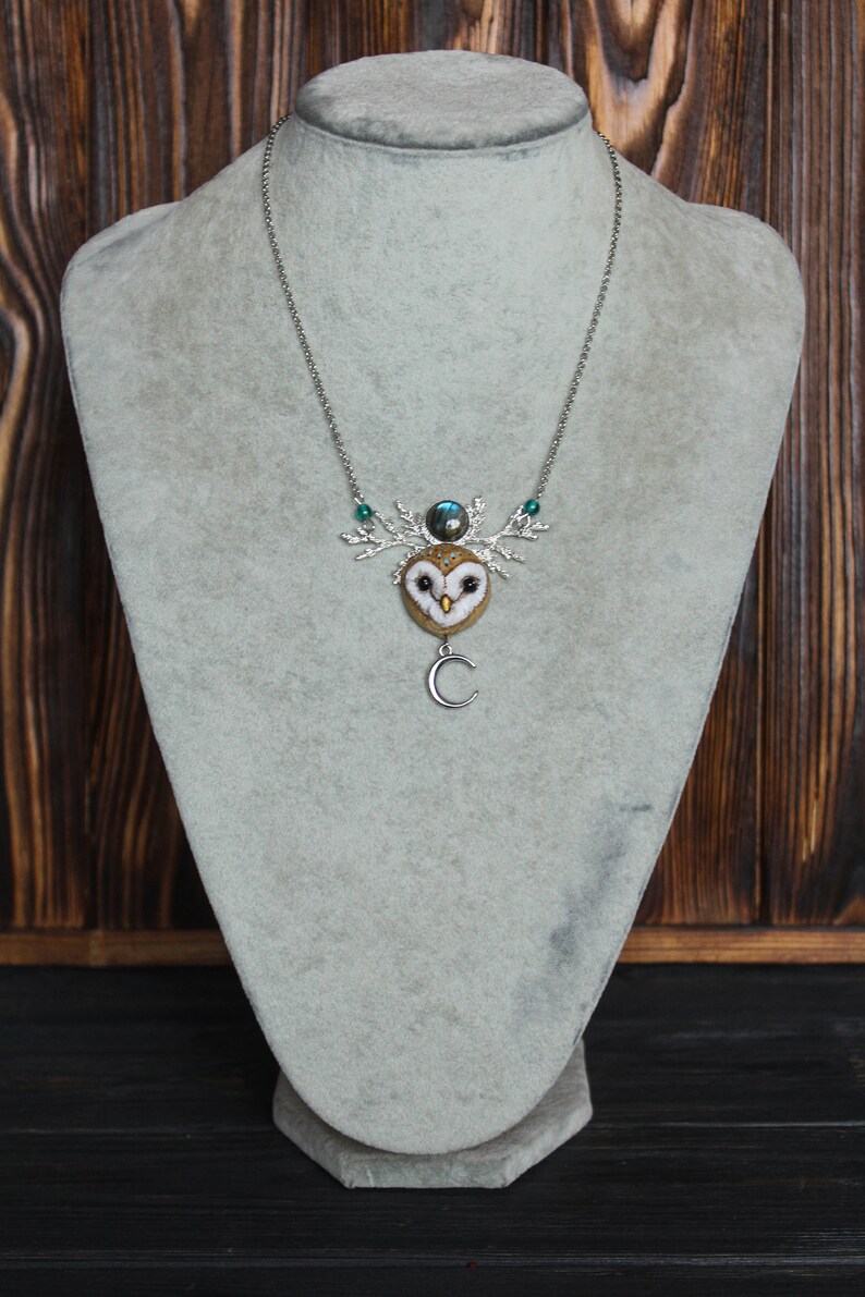 May include: A silver chain necklace with a felt owl pendant. The owl has a brown and white pattern and a green gemstone on its head. The pendant is attached to a silver branch with a crescent moon charm.