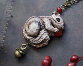May include: A handcrafted pendant necklace featuring a detailed squirrel figurine holding a red berry. The squirrel is painted in shades of brown and gray, with a small acorn charm hanging below. The necklace chain is antique brass.