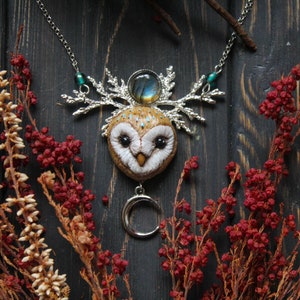 May include: A silver necklace with a brown owl pendant, a silver crescent moon charm, and a blue-green gemstone. The owl has a white face with black eyes and a brown body with white spots. The necklace is on a dark wooden background with red and brown dried flowers.