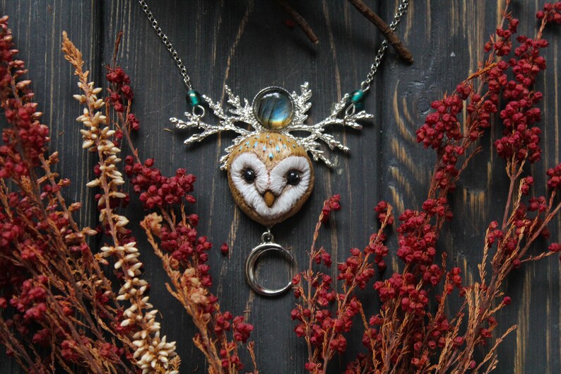 May include: A silver necklace with a brown owl pendant, a silver crescent moon charm, and a blue-green gemstone. The owl has a white face with black eyes and a brown body with white spots. The necklace is on a dark wooden background with red and brown dried flowers.