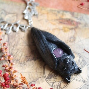 May include: A handcrafted black bat pendant with a silver chain. The bat features a purple gemstone and detailed texture. The pendant is displayed on a vintage map background, with red and orange floral accents.