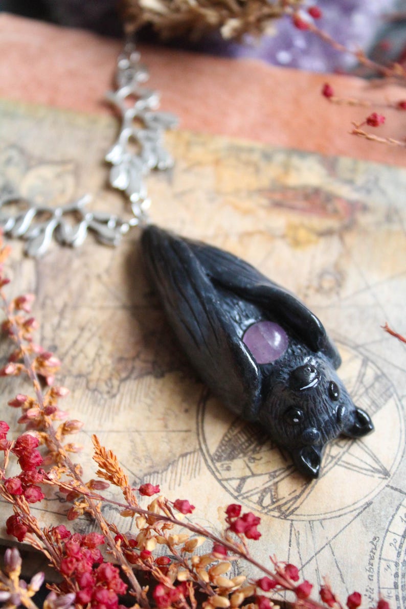 May include: A handcrafted black bat pendant with a silver chain. The bat features a purple gemstone and detailed texture. The pendant is displayed on a vintage map background, with red and orange floral accents.