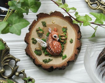 May include: A whimsical wooden slice wall hanging featuring a fawn with fairy wings. The fawn is brown with white spots, surrounded by green moss and miniature mushrooms. A crescent moon and gold accents add detail to this woodland-themed decor.
