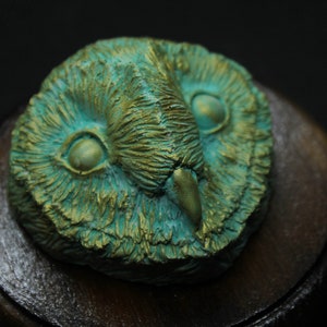 May include: A detailed, close-up of an owl head sculpture. The owl is rendered in shades of teal and gold, with intricate feather detailing. The sculpture is mounted on a dark wooden base, creating a striking contrast against a black background.