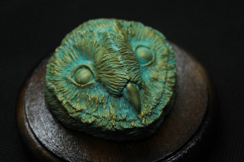 May include: A detailed, close-up of an owl head sculpture. The owl is rendered in shades of teal and gold, with intricate feather detailing. The sculpture is mounted on a dark wooden base, creating a striking contrast against a black background.