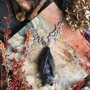 May include: A dark gray bat pendant with a purple gemstone belly, suspended from a silver-toned leaf-design chain. The pendant rests on a vintage map, surrounded by red and brown floral accents and amethyst crystals.