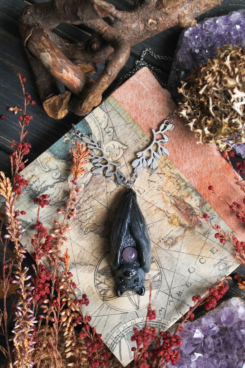 May include: A dark gray bat pendant with a purple gemstone belly, suspended from a silver-toned leaf-design chain. The pendant rests on a vintage map, surrounded by red and brown floral accents and amethyst crystals.