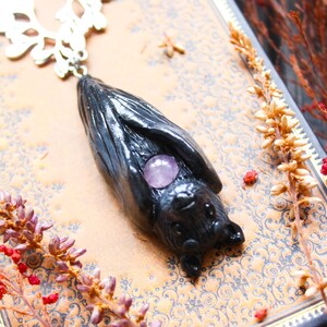 May include: A black bat pendant with a silver-colored filigree chain. The bat has a purple gemstone in the center. The pendant is lying on a patterned book with dried flowers and berries.