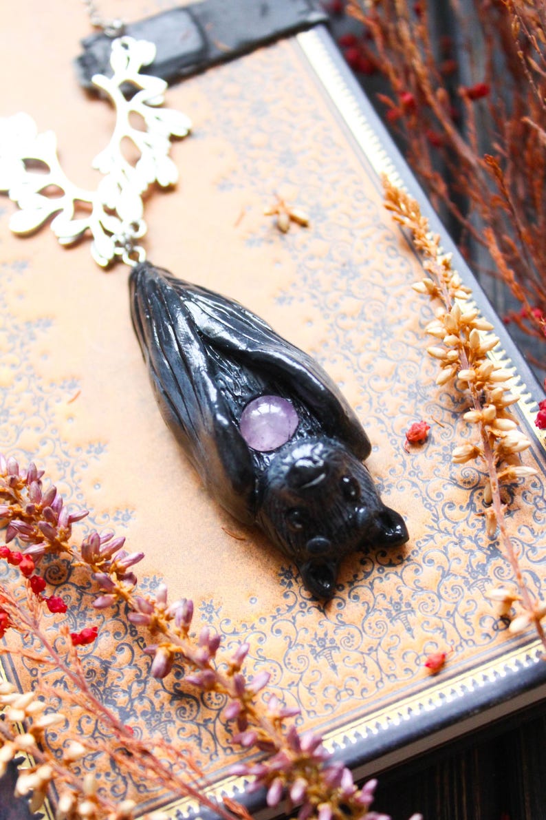 May include: A black bat pendant with a silver-colored filigree chain. The bat has a purple gemstone in the center. The pendant is lying on a patterned book with dried flowers and berries.