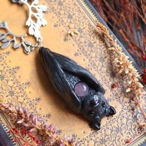 May include: A handcrafted necklace featuring a dark gray bat pendant with a purple gemstone. The bat hangs from a silver chain with a decorative branch-like element. The necklace rests on a vintage book with floral patterns.