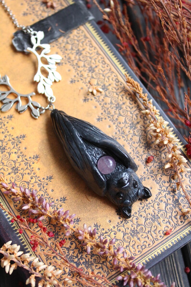 May include: A handcrafted necklace featuring a dark gray bat pendant with a purple gemstone. The bat hangs from a silver chain with a decorative branch-like element. The necklace rests on a vintage book with floral patterns.
